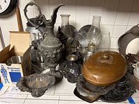 Photo showing assorted vintage metal and glass items including ornate metal pitcher, tarnished silver serving platter and dishes, pewter mug, hurricane lamps and chimneys
