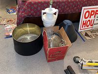 White ceramic lamp base with lamp socket, two black lamp shades, and cardboard box with glass hanging lamp parts on carpeted floor in front of a red patterned bedspread.