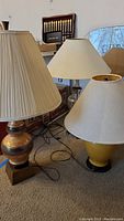 Three lamps with different shades, bases ceramic and glass. One has pleated beige shade with ornate bronze and gold ceramic base on square bottom. Second has simple beige shade and glass base with brass neck. Third has white umbrella-shaped shade and yellow ceramic base with black bottom. Lamps dusty and untested.