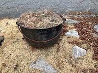 Close-up front side image of cast iron cauldron planter showing rusted surface and filled with dried grass and leaves.