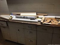 Rolled posters and maps stacked on a cabinet surface, showing multiple tubes and rolled papers in worn condition.