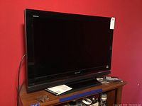 Sony Bravia 32-inch flat screen TV with black bezel on wooden stand with two remote controls and instruction manual.