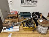 Overview photo of lot on table showing two plastic tool totes, a green metal toolbox, flashlight, spools of white rope and cable, and assorted hand tools including measuring sticks and scissors.
