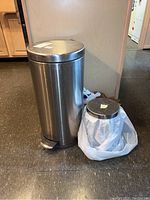 Photo of two stainless steel cylindrical trash cans with foot pedals, one large and one small, placed on floor against wall.
