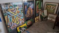 Multiple framed paintings and posters leaning against a wall, showing different themes: abstract, figurative, pastoral. Sizes vary with frames.