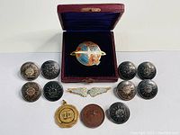 Entire lot displayed including leather jewelry box, colorful globe pin, sweetheart pilot wings, bronze medallions, and military buttons arranged on white background.
