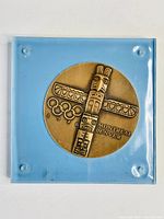 Front view of bronze medallion displaying Indigenous totem pole style figure with Olympic rings and '1976 Montreal Denver' text in blue acrylic block.