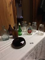Full view of assorted vintage glass bottles and jars arranged on a white cloth. Includes clear, amber, and green glass bottles with varied neck styles and containers including a Log Cabin Syrup bottle.