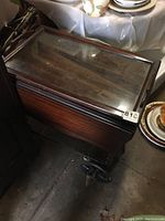 Wooden butler tray with a glass top resting on dark wooden rolling tea cart. The tray shows surface marring and the setup is positioned in a storage or display area.