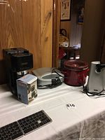 Wide angle photo showing all four appliances on a white table, including DeLonghi espresso machine, Cuisinart sandwich maker, Crock Pot, and Oster can opener.