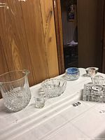 Photo shows all items included in the lot arranged on a striped white cloth table covering. Includes a large crystal ice bucket, Waterford crystal clock with chip, small clear creamer, two crystal bowls, pair of crystal votives, and a blue and clear decorative bowl.