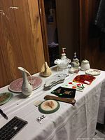 Photo of table with assorted household decor items including ceramic pitchers, plates, soap dispenser, glass shakers, red glass panel, wooden hairbrush.