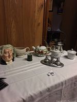 View of entire tea set on white tablecloth including large face-shaped mug, tea kettle, teapot with strawberry design, sugar bowl, creamer, gravy boat, and salt and pepper shakers on silver stand.