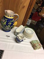 Wide view of large blue floral pitcher, 4 white corn trays stacked on each other, 4 blue and green saucers stacked on each other, small blue and white pitcher, tied set of 4 green coasters on table