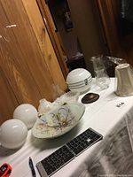 Photo showing two white glass globes, decorative floral glass lamp shade, several glass lamp chimneys, and dark metal ring fixture on table