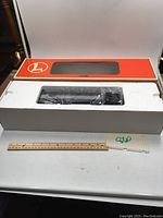 Top-down photo of the Lionel Heavyweight Baggage Car unopened in its white molded foam packaging inside its open orange Lionel branded box, resting on a table with ruler for scale.