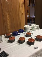 Set of eight small handled pots arranged on a table; includes 2 blue pots with lids and spouts and 6 brown Guernsey Ware pots with metal holders and lids.