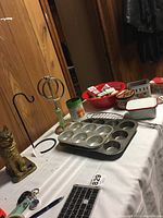 Photo showing brass cat figurine, black metal towel rack, muffin pans, red Zak bowl, and spice containers on a white table.