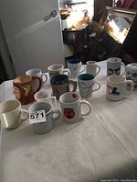 Wide shot of 12 assorted coffee and tea mugs on white tablecloth, featuring various colors, designs, and styles.