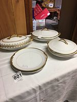 Five cream and gold china pieces on white tablecloth including three covered dishes and two platters with handles.