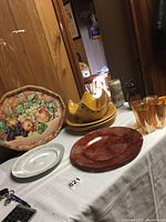 Wide view photo showing assorted pottery and glassware including large Pottery Barn plates, orange mug, set of four orange tinted glasses, fluted bowl, metal tray with fruit design, and other pieces on a table.