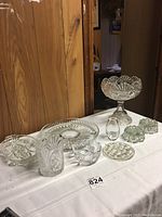 Full view of the set of glass decorative items arranged on a table, showing platter, bowls, vases, frog, compote, and candleholders.