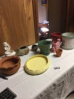 Photo of assorted pots and decorative items on a table, showing various shapes, sizes, and colors including green, yellow, white, pink, terracotta, and brown.