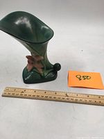 Green glazed Roseville pottery lily-shaped vase with a pink flower detail, small size with a chip under the lip, placed next to a ruler for scale.