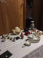 Wide view of two teapots, miniature tea set, heart-shaped cheese molds, lacquer coasters, salt and pepper shakers, bell, and candles on a table with wood-paneled background.