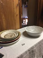 Photo showing stacked large platters and bowls on a white tablecloth. The plates vary in design and size with decorative rims, and a clear glass bowl with a shell motif is visible to the right.