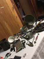 Photo showing various vintage metal kitchen items spread on table including sifters, graters, ice cream scoops, nut crackers, mallet and more.
