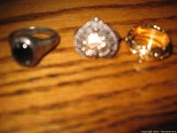 Three sterling silver rings shown side by side on wood surface. Included are a black stone ring, a heart-shaped cluster stone ring, and a decorative band with cutouts and gold toned inside.