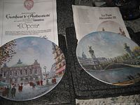 Photo of two collectible plates showing Paris landmarks 'L'Opera' and 'Le Pont Alexandre III' with their certificates of authenticity, displayed over protective cases.
