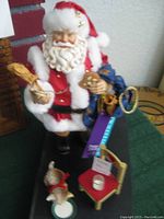 Santa Claus figurine standing on a green surface with a blue sack, holding a cookie and a brown sack, a small gray kitten on a round white base near a miniature gold chair with a cookie and milk glass.