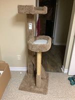 Full view of tan carpeted cat tree with two resting platforms and sisal scratching post, on carpeted floor with cardboard box and weights nearby.