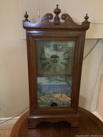 Full front view showing the wooden case, the painted clock face with Roman numerals, decorative floral corners, and the lower scenic panel with a coastal landscape painting.