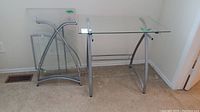 Partially disassembled metal frame and glass tops for an L-shaped desk, showing frame sections and glass panels.