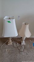 Two marble base lamps side by side on carpet. Larger lamp on left has rectangular white shade and smaller lamp on right has cream bell shade.