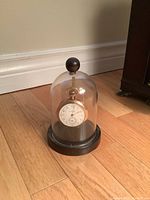 Gold tone pocket watch displayed on black base with clear plastic dome cover.
