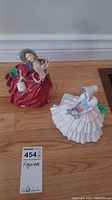 Two Royal Doulton porcelain figurines on hardwood floor, one in red dress, one in white dress.