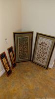 Full view of two large framed silk art pieces and two smaller framed metal art pieces, arranged against corner walls on floor.