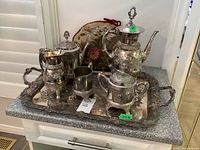 Full set of silverplate and metal tea service set displayed on ornate tray with fabric floral pillow background
