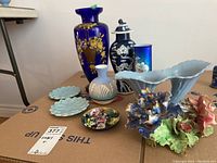 Full view of the lot showing all items on cardboard including cobalt blue vases, pale blue plates, floral figurines, and trinket dish.