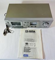 Vintage Akai CS-M01A stereo cassette deck with control panel, cassette compartment, and power cord alongside original manual.
