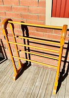 Full view of an oak quilt rack with four horizontal bars, standing on a wooden floor against a brick wall in daylight.
