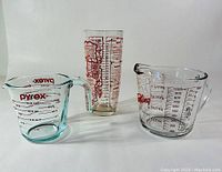 Front view of three glass measuring cups and mixer with red printed markings and cocktail recipes.