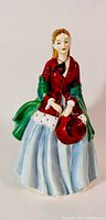 Front view of the porcelain figurine showing the lady in a red jacket with green shawl and light blue skirt, holding a red hat in her hands.