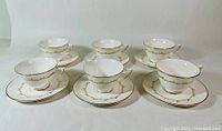 Front view of 6 white fine bone china cups with matching saucers, decorated with gold trim and intricate gold design around each cup and saucer edge.