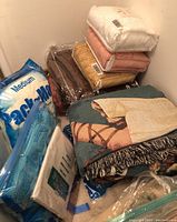 Photo showing a stack of bed linens including packaged bed sheets in white, blue, and other colors, a medium size mattress protector, and two folded blankets with patterns laying on top.