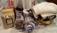 Overview of all blankets and throws: three sealed Comfort Collection blankets, three cylindrical Shavel rolls, pile of folded throws